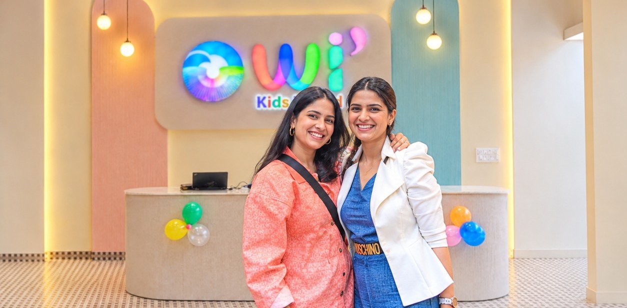 Two smiling friends hugging in front of the glowing OWI Kids World logo at the reception desk